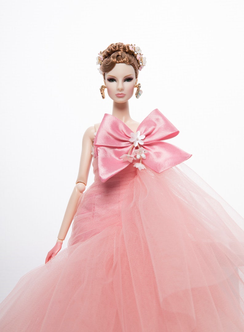 May include: A pink tulle gown with a large pink satin bow and white flower embellishments. The doll is wearing a floral crown and gold earrings.