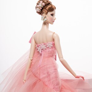 May include: A pink tulle ball gown with a floral design and a bow on the back. The doll is wearing a butterfly earring.