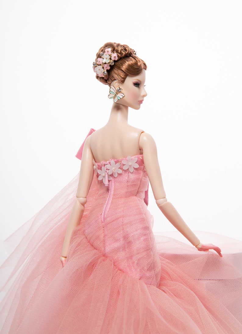 May include: A pink tulle ball gown with a floral design and a bow on the back. The doll is wearing a butterfly earring.