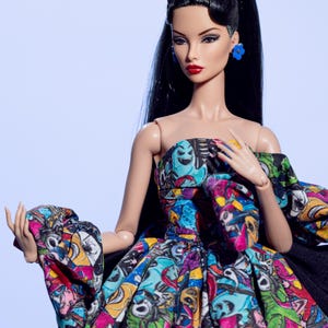 May include: A fashion doll with long black hair, red lipstick, and blue flower earrings. The doll wears a strapless dress with a colorful, cartoon-like print and a black cape. A light blue bow adorns the doll's hair.