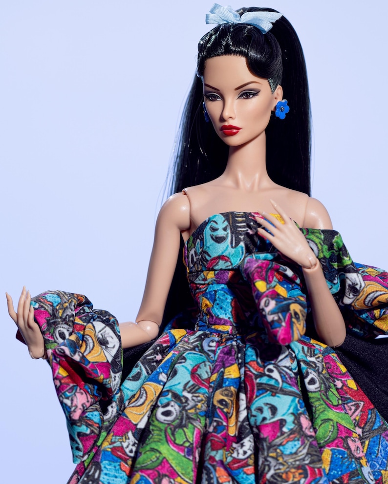 May include: A fashion doll with long black hair, red lipstick, and blue flower earrings. The doll wears a strapless dress with a colorful, cartoon-like print and a black cape. A light blue bow adorns the doll's hair.