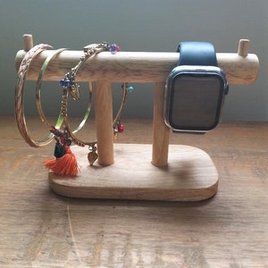 May include: A wooden stand with two arms for holding jewellery and a watch. The stand is made of light brown wood and has a natural finish. There are three gold bracelets with colourful beads and tassels hanging from the arms. A black and silver smartwatch is resting on the top arm.