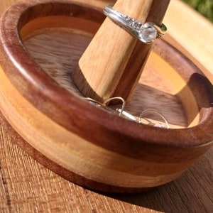 May include: A silver ring with a clear stone and small accent stones is displayed on a wooden ring holder. The ring holder is inside a wooden bowl with a layered design. The bowl and ring holder are on a wooden surface.