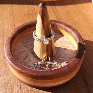 May include: A wooden ring holder with a silver ring and a pair of earrings. The holder is a light brown cone shape, set in a round, multi-toned wooden dish. The dish has a dark brown rim and a lighter wood interior.