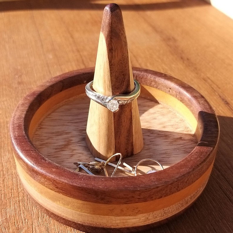 May include: A handcrafted wooden ring holder with a conical post and a round base. The holder features a two-tone design with a dark brown and light brown wood grain. A diamond ring is displayed on the post, with other rings in the base.