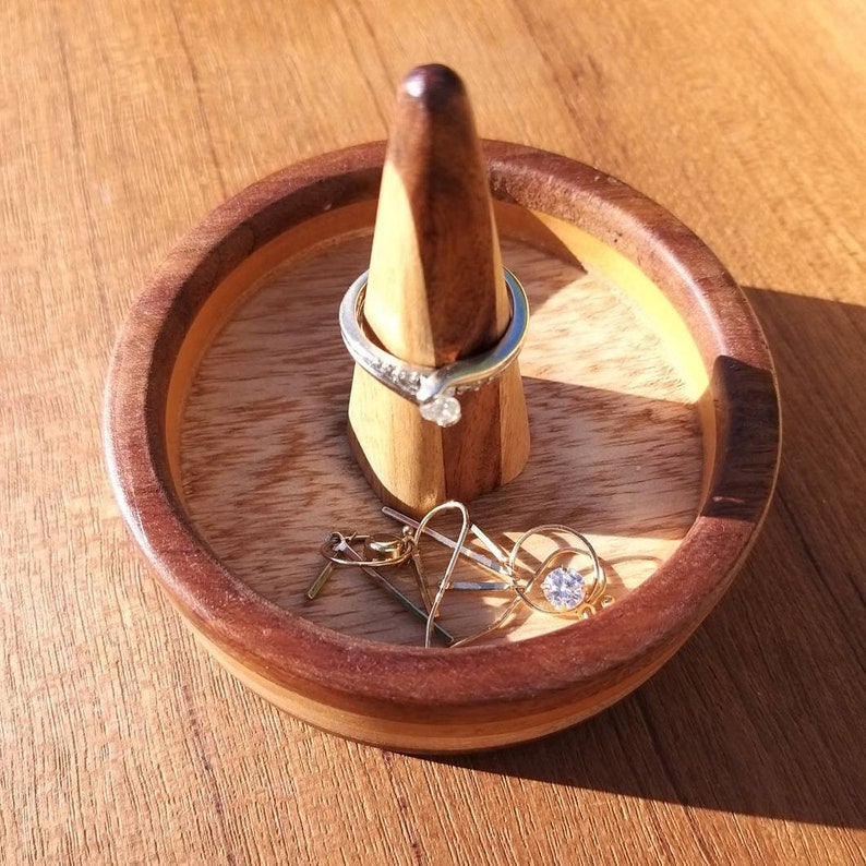 May include: A round wooden jewelry holder with a dark brown rim and a light brown interior. A silver ring with a clear stone sits on a wooden cone in the center. Gold earrings with clear stones are also in the holder.