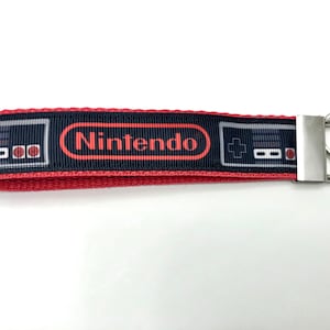 May include: A black and red key chain with a Nintendo logo and a retro video game controller design. The key chain has a silver metal key ring.