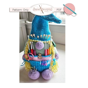 Op de afbeelding: Een gehaakte gnoomvormige garenorganizer met een blauwe muts, een groen lichaam en paarse voeten. De gnoom heeft een zakje aan de voorkant om scharen en andere kleine spullen in op te bergen. De gnoom is gevuld met breinaalden, haaknaalden en andere benodigdheden.