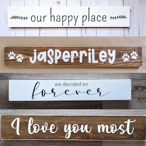 May include: Four rectangular wooden signs with text. The top sign is white with black text reading 'our happy place'. The second is dark brown with white text 'JASPERRILEY' and paw prints. The third is white with black text 'we decided on forever'. The bottom is dark brown with white text 'I love you most'.