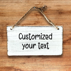 以下が含まれることがあります： 中央に黒い文字で「Customized your text」と印刷された、使い古した質感の白い長方形の木製看板。看板は、板の上部の角に取り付けられた自然な茶色の麻紐で吊り下げられています。