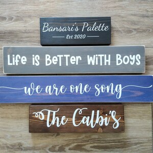 May include: Four rectangular wooden signs with white lettering. The top sign is black with 'Bansari's Palette Est 2020'. The second is gray with 'Life is BeTTer WITH BOYS'. The third is blue with 'we are one song'. The bottom is brown with 'The Calbi's'.