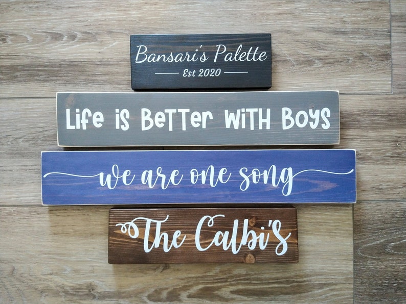 May include: Four rectangular wooden signs with white lettering. The top sign is black with 'Bansari's Palette Est 2020'. The second is gray with 'Life is BeTTer WITH BOYS'. The third is blue with 'we are one song'. The bottom is brown with 'The Calbi's'.
