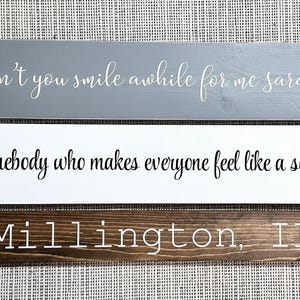 May include: Three rectangular wooden signs. The top sign is gray with white cursive text reading won't you smile awhile for me sara. The middle sign is white with black cursive text reading Be somebody who makes everyone feel like a somebody. The bottom sign is dark brown with white text reading Millington, IL.