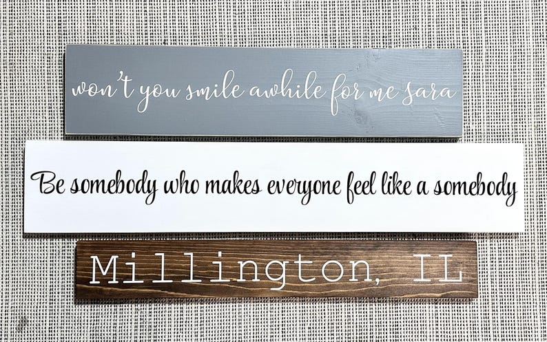 May include: Three rectangular wooden signs. The top sign is gray with white cursive text reading won't you smile awhile for me sara. The middle sign is white with black cursive text reading Be somebody who makes everyone feel like a somebody. The bottom sign is dark brown with white text reading Millington, IL.