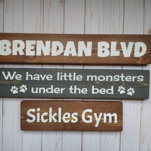 May include: Three rectangular wooden signs with white lettering. The top sign reads BRENDAN BLVD. The middle gray sign reads We have little monsters under the bed with paw prints. The bottom sign reads Sickles Gym. All signs feature visible wood grain.