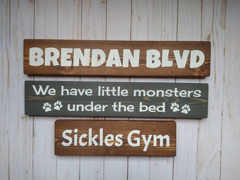 May include: Three rectangular wooden signs with white lettering. The top sign reads BRENDAN BLVD. The middle gray sign reads We have little monsters under the bed with paw prints. The bottom sign reads Sickles Gym. All signs feature visible wood grain.