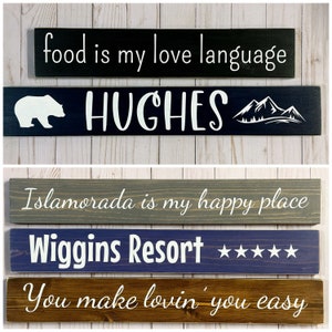 May include: Five rectangular wooden signs with white text and graphics. The top sign is black with "food is my love language". The second is dark blue with a bear, "HUGHES", and mountains. The third is gray with "Islamorada is my happy place". The fourth is blue with "Wiggins Resort" and five stars. The bottom is brown with "You make lovin' you easy".