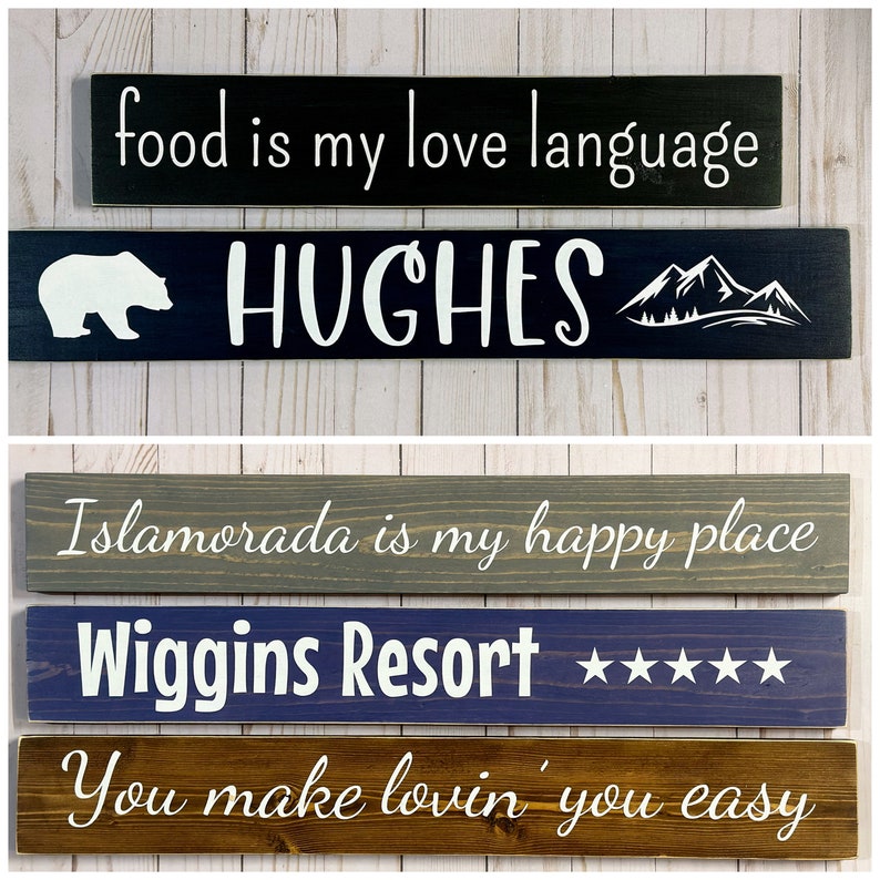 May include: Five rectangular wooden signs with white text and graphics. The top sign is black with "food is my love language". The second is dark blue with a bear, "HUGHES", and mountains. The third is gray with "Islamorada is my happy place". The fourth is blue with "Wiggins Resort" and five stars. The bottom is brown with "You make lovin' you easy".