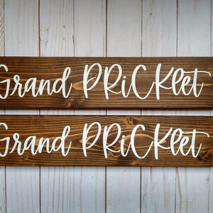 May include: Two rectangular wooden signs with a dark brown stained finish and visible wood grain. Each sign features the white text "Grand PRiCKLET" in a cursive script font. The signs are positioned horizontally, one above the other.