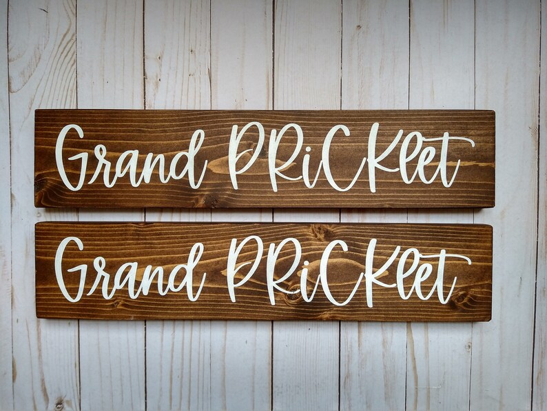 May include: Two rectangular wooden signs with a dark brown stained finish and visible wood grain. Each sign features the white text "Grand PRiCKLET" in a cursive script font. The signs are positioned horizontally, one above the other.