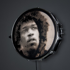 Può includere: Un tamburo nero e argento con un ritratto di un viso umano ritagliato dalla pelle del tamburo. Il viso è illuminato da dietro, creando un effetto luminoso.