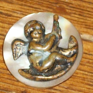 Può includere: Un bottone vintage con un angelo cherubino con ali, seduto su un'altalena. Il bottone è in metallo e ha una finitura madreperlacea.