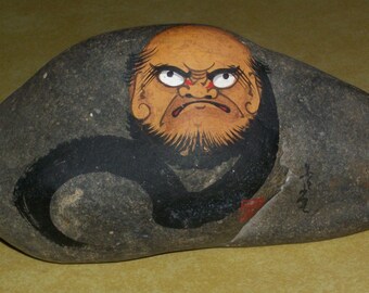 May include: A gray stone with a black and orange painted image of a scowling face with red eyes. The face is surrounded by a black swirl.