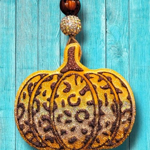 May include: A gold glitter pumpkin ornament with a brown leopard print design. The ornament is hanging from a string with brown and clear beads.