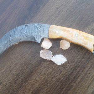 May include: A curved, Damascus steel knife with a bone handle and a ring on the end. The knife is resting on a wooden surface with pink Himalayan salt rocks.