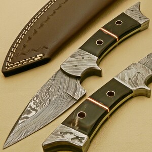 May include: Two Damascus steel knives with black and brown handles. The blades have a wavy pattern and the handles have a copper inlay.
