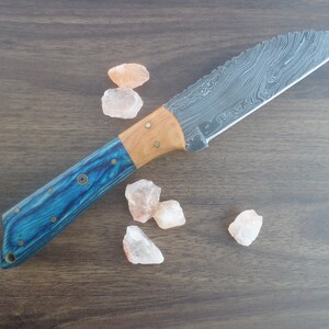 Puede incluir: Un cuchillo con mango azul y madera con una hoja de acero Damasco. El cuchillo está sobre una superficie de madera con varios cristales pequeños de color rosa.