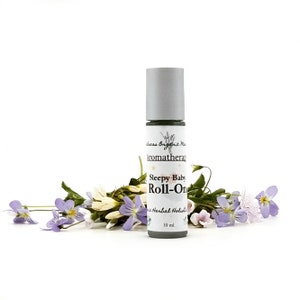 May include: A white roll-on bottle with a green label that says "Sleepy Baby Roll-On" and "Herbal Holistic" with a lavender graphic. The bottle is 10 ml. and is surrounded by purple and white flowers.