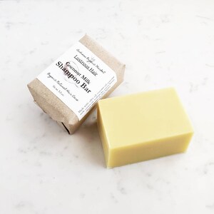 May include: A light yellow, rectangular shampoo bar next to a wrapped bar with a label. The label reads "Lustrous Hair Coconut Milk Shampoo Bar" and "Organic Natural Hair Care". The unwrapped bar is approximately 3.5 inches long and 2 inches wide.