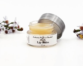 May include: A small, open glass jar of vanilla lip balm with a silver lid. The label reads "Andreas Organic Market Vanilla Lip Balm .25 oz." The balm is a light yellow color. White flowers are on either side of the jar.