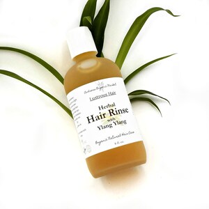 May include: A clear glass bottle of herbal hair rinse with a white cap, labeled "Lustrous Hair" and "Herbal Hair Rinse with Ylang Ylang." The bottle contains a golden liquid and is 8 fl oz. Green leaves are in the background.