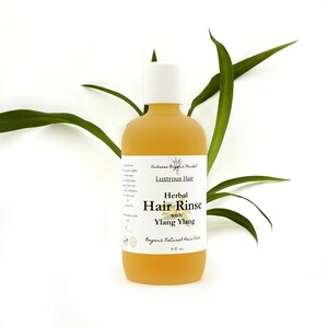 May include: A yellow glass bottle with a white cap, labeled "Lustrous Hair Herbal Hair Rinse with Ylang Ylang." The bottle is 8 fl. oz. and is set against a white background with green leaves.