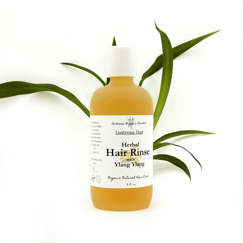 May include: A yellow glass bottle with a white cap, labeled "Lustrous Hair Herbal Hair Rinse with Ylang Ylang." The bottle is 8 fl. oz. and is set against a white background with green leaves.