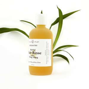 May include: A frosted glass bottle of herbal hair rinse with a white cap. The label reads "Lustrous Hair Herbal Hair Rinse with Ylang Ylang." The bottle contains a yellow liquid and is 8 fl oz. Green leaves are in the background.