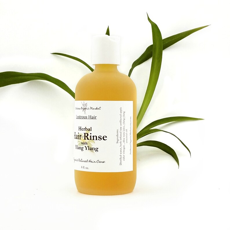 May include: A frosted glass bottle of herbal hair rinse with a white cap. The label reads "Lustrous Hair Herbal Hair Rinse with Ylang Ylang." The bottle contains a yellow liquid and is 8 fl oz. Green leaves are in the background.