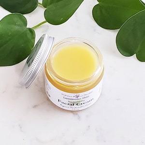 May include: A small glass jar of yellow facial cream with a silver lid, labeled "Luminous Skin Facial Cream." The jar is open, revealing the cream's texture. Green leaves are in the background, creating a natural aesthetic.