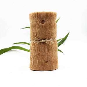 May include: A cylindrical, textured, light brown candle with a rustic appearance. The candle is wrapped with twine and tied in a bow. Green leaves are visible in the background, set against a white backdrop. The candle has a natural, organic aesthetic.