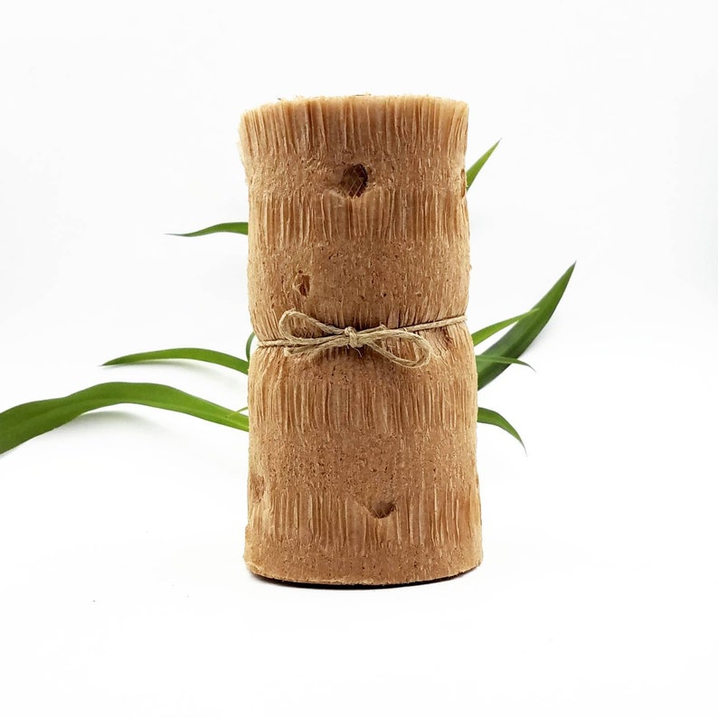 May include: A cylindrical, textured, light brown candle with a rustic appearance. The candle is wrapped with twine and tied in a bow. Green leaves are visible in the background, set against a white backdrop. The candle has a natural, organic aesthetic.