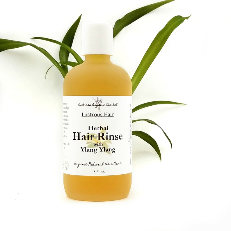 May include: A bottle of herbal hair rinse with a white cap and a yellow liquid. The label reads "Lustrous Hair" and "Herbal Hair Rinse with Ylang Ylang." The bottle is 8 fl. oz. and is set against a white background with green leaves.