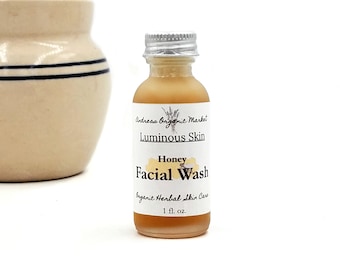 May include: A small glass bottle of honey-colored facial wash with a silver cap. The label reads "Luminous Skin Honey Facial Wash" from Andreas Organic Market. The bottle contains 1 fl. oz. of product. A ceramic jar with blue stripes is in the background.