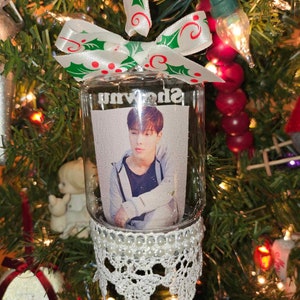 May include: A clear glass ornament with a white lace trim and a white pearl border. Inside the ornament is a photo of a person with brown hair. The ornament is decorated with a white and green bow with red polka dots. The text "Shownu" is printed on the ornament.