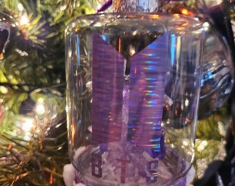 Op de afbeelding: Een transparante glazen kerstbal met een paars, iriserend BTS-logo erin. De bal is versierd met witte pompoenen en een paars lint.