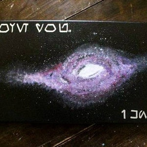 Peut inclure: Une peinture sur toile noire avec un motif de galaxie violet et blanc. Le texte "1 JOYVT VOUL." est en haut de la toile et "1 JΛOO." est en bas.