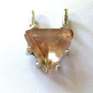 May include: A triangular pendant with a faceted, peach-colored gemstone. The gemstone is set in a silver-colored metal setting with two loops at the top for a chain. The pendant has a pointed bottom and is displayed against a white background.