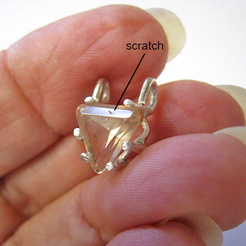 May include: A triangular, light-colored gemstone pendant with a silver setting. The gemstone has a visible scratch. The pendant has two loops at the top for a chain and small decorative prongs around the edges.
