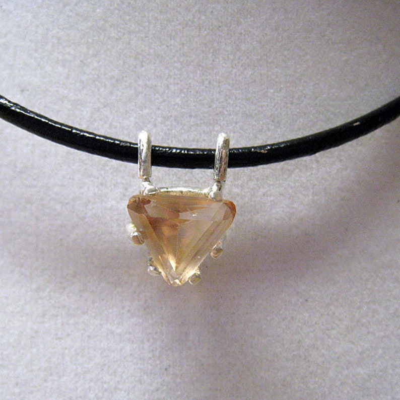 May include: A pendant necklace featuring a triangular, light-orange gemstone set in a silver metal frame. The pendant hangs from a black, leather-like cord. The gemstone has a faceted surface, catching the light.